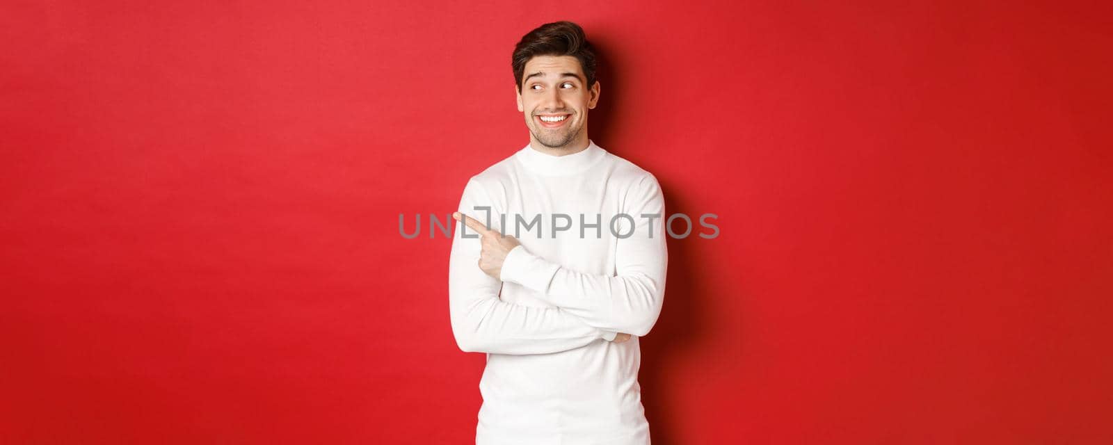 Concept of winter holidays, christmas and lifestyle. Excited handsome man in white sweater, smiling, looking and pointing finger left at promo offer, standing over red background.