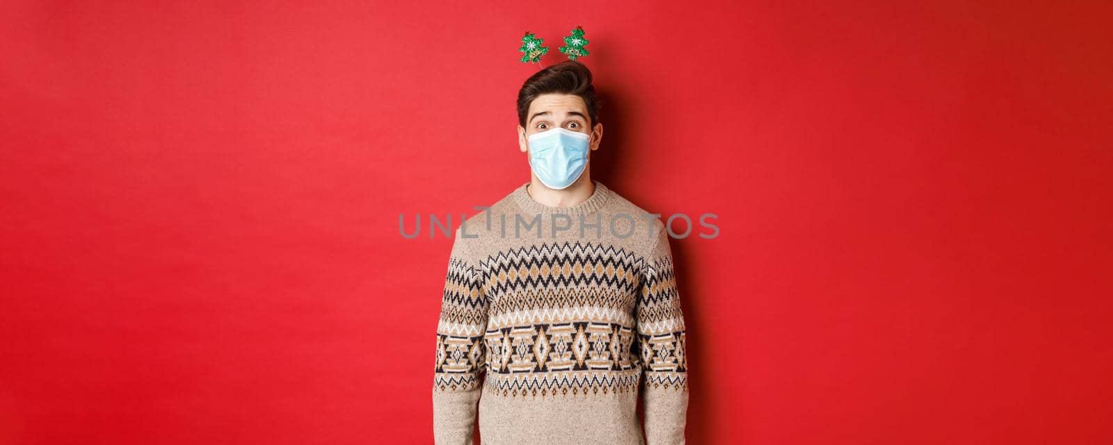 Concept of christmas, covid-19 and social distancing. Cheerful young man in medical mask and new year clothing, celebrating holidays during pandemic, standing over red background.
