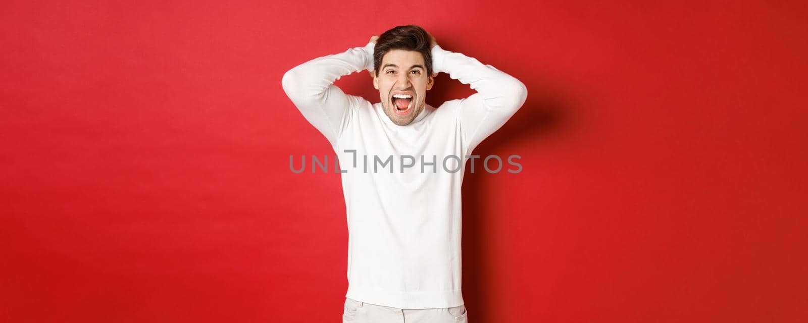 Portrait of frustrated man in white sweater, shouting in anger, holding hands on head and grimacing, feeling distressed, standing over red background.
