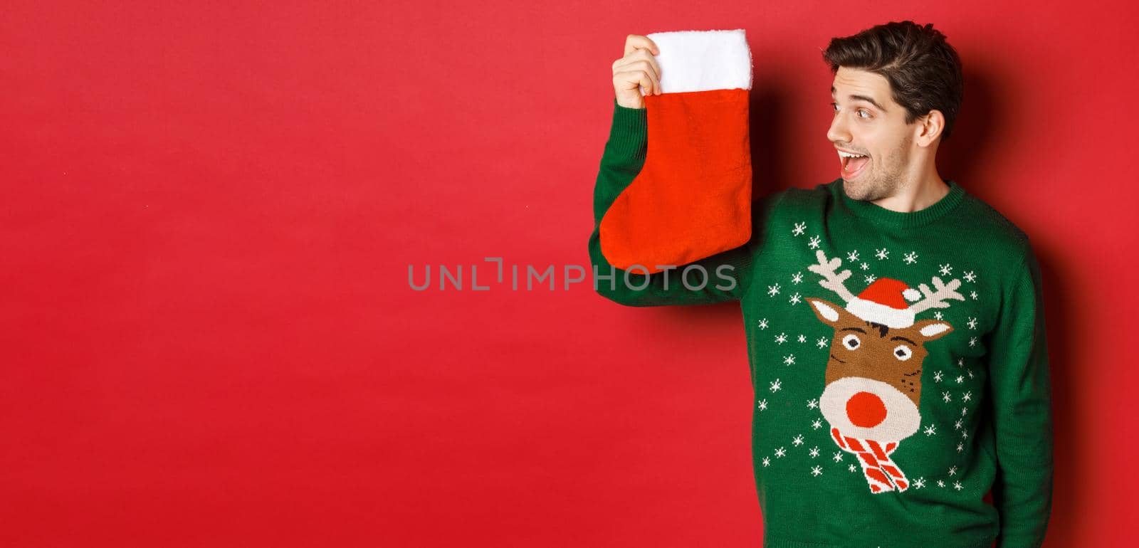 Image of surprised and amused man in green sweater, looking at christmas stocking with presents and smiling, standing over red background by Benzoix