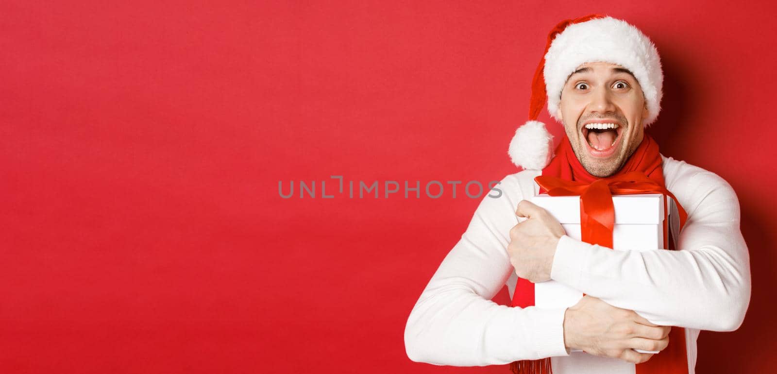 Concept of winter holidays, christmas and lifestyle. Close-up of happy man in santa hat receiving present, looking happy and hugging gift box, standing over red background.