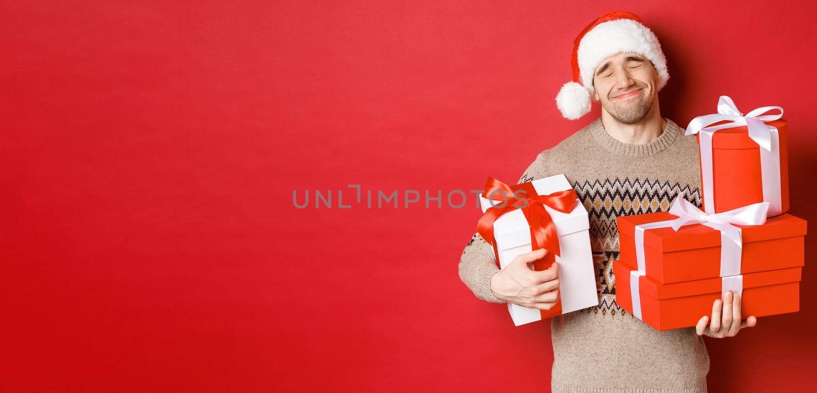 Concept of winter holidays, new year and celebration. Portrait of lovely smiling man receiving pile of presents, holding gifts and being touched with surprise, standing over red background grateful.