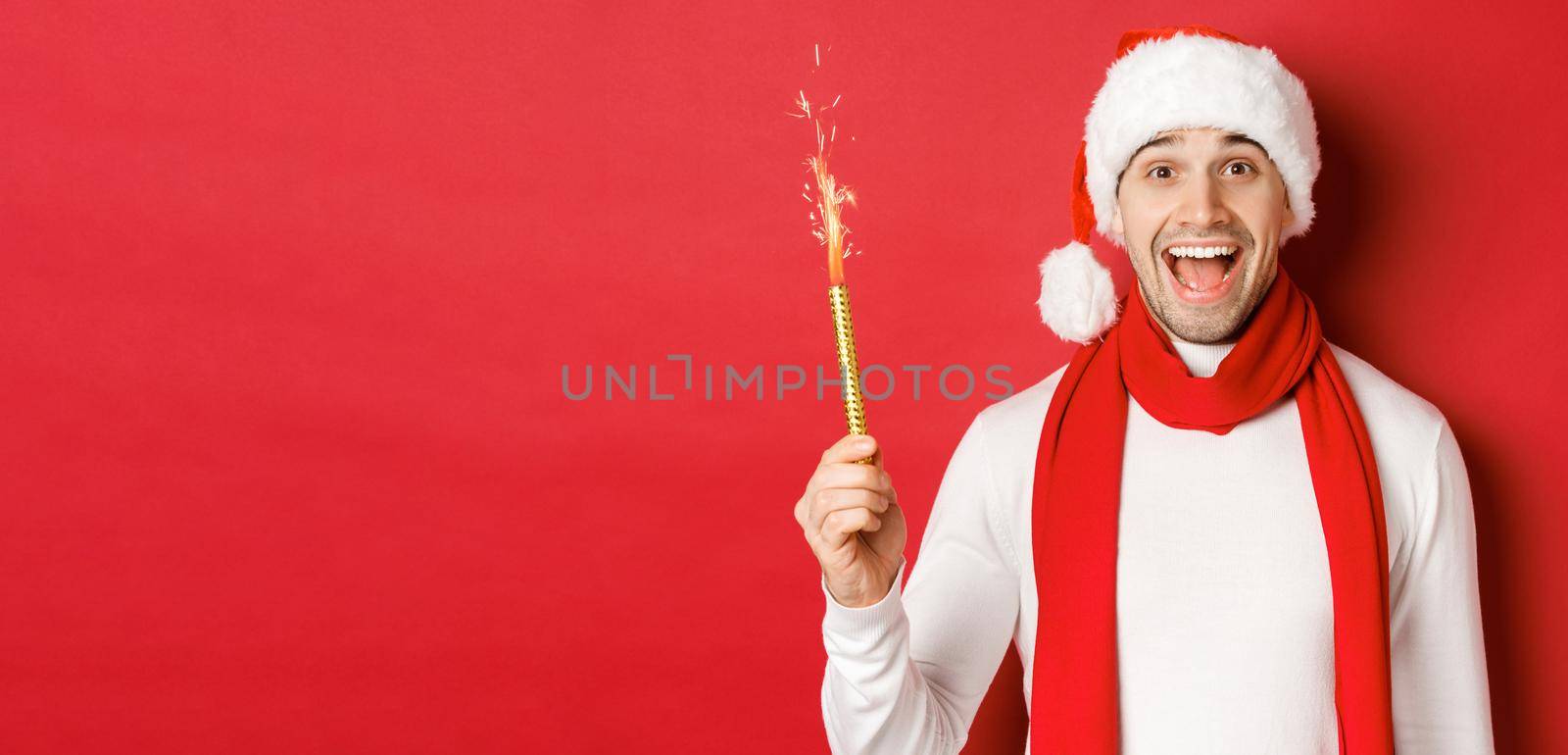 Concept of christmas, winter holidays and celebration. Handsome man celebrating new year and having fun, holding sparkler and smiling, wearing santa hat, standing over red background.