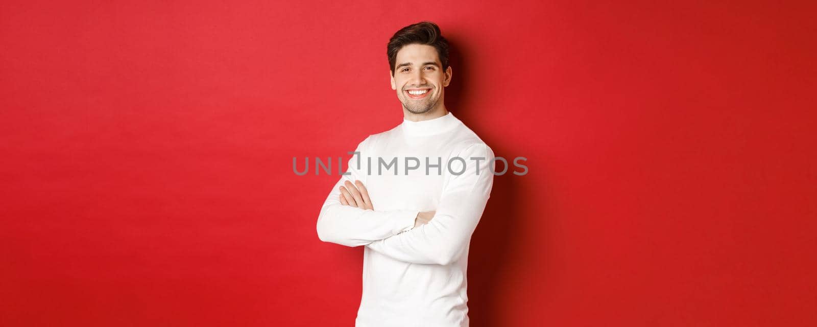 Concept of winter holidays, christmas and lifestyle. Portrait of confident attractive guy in white sweater, smiling with hands crossed on chest, standing over red background.