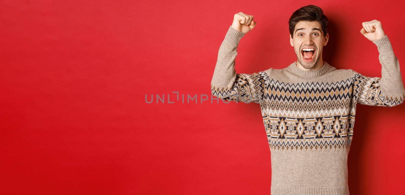 Image of happy handsome guy rejoicing on christmas eve, raising hands up and shouting for joy yes, achieve new year goal, standing over red background in sweater.