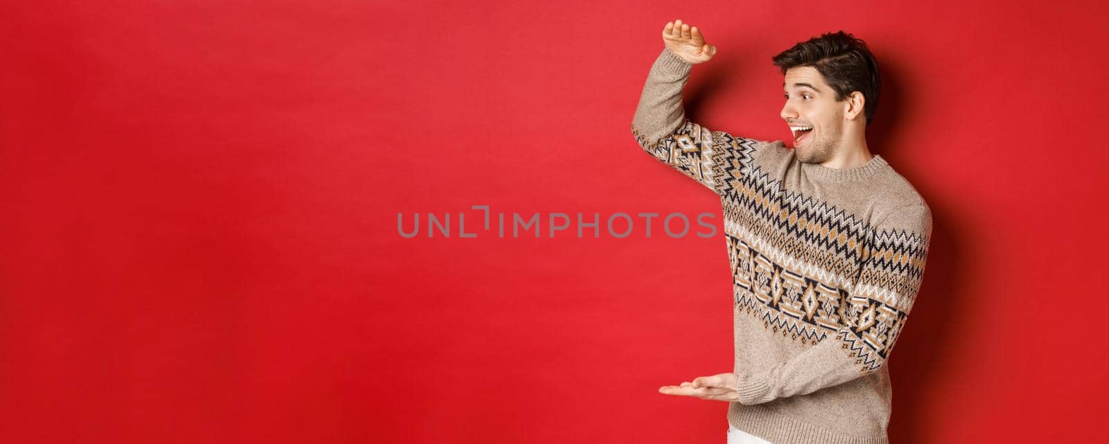 Portrait of happy young man in christmas sweater, showing large present, smiling and looking amazed at cool gift, standing over red background by Benzoix