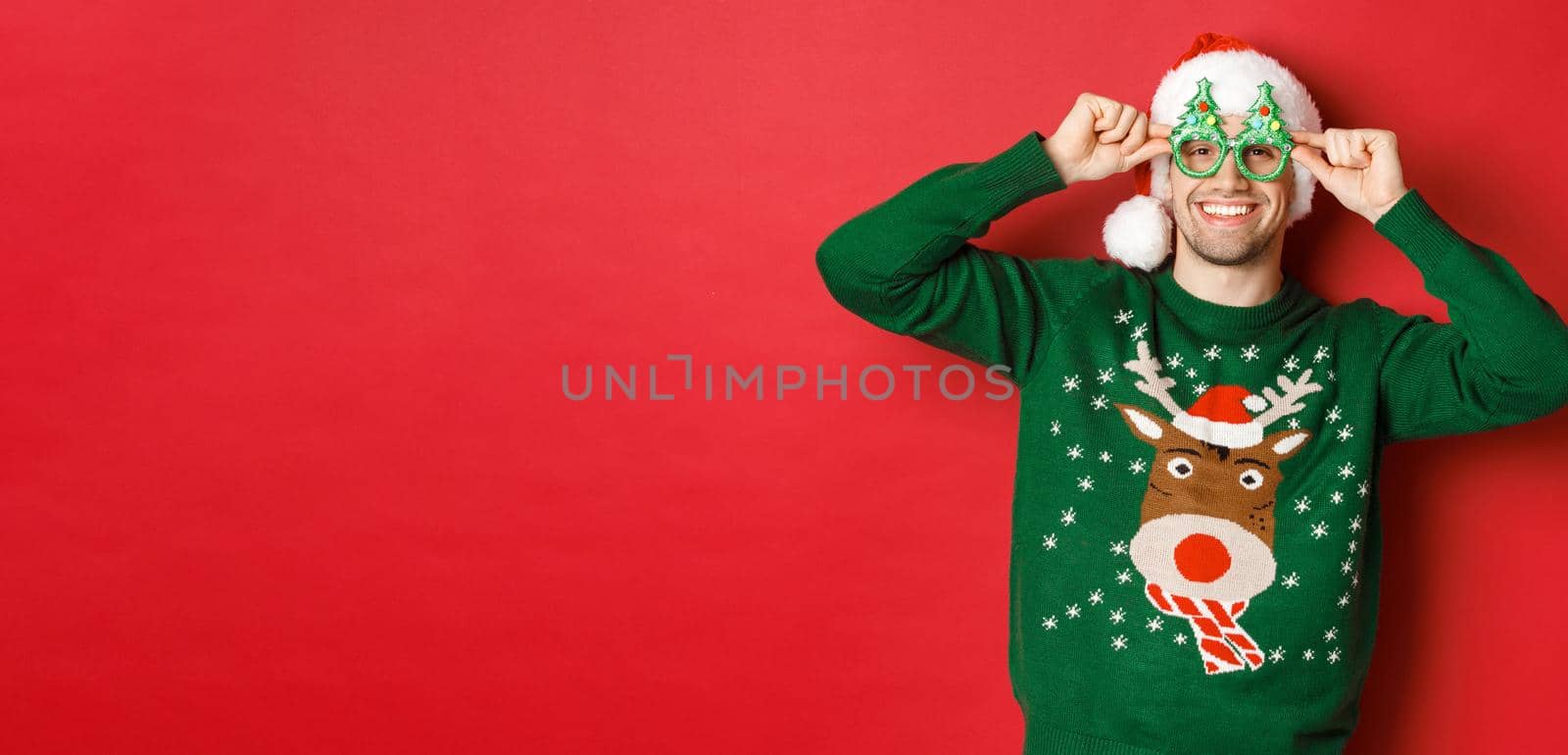 Portrait of attractive smiling man in santa hat, party glasses and sweater, celebrating new year holidays, standing against red background.