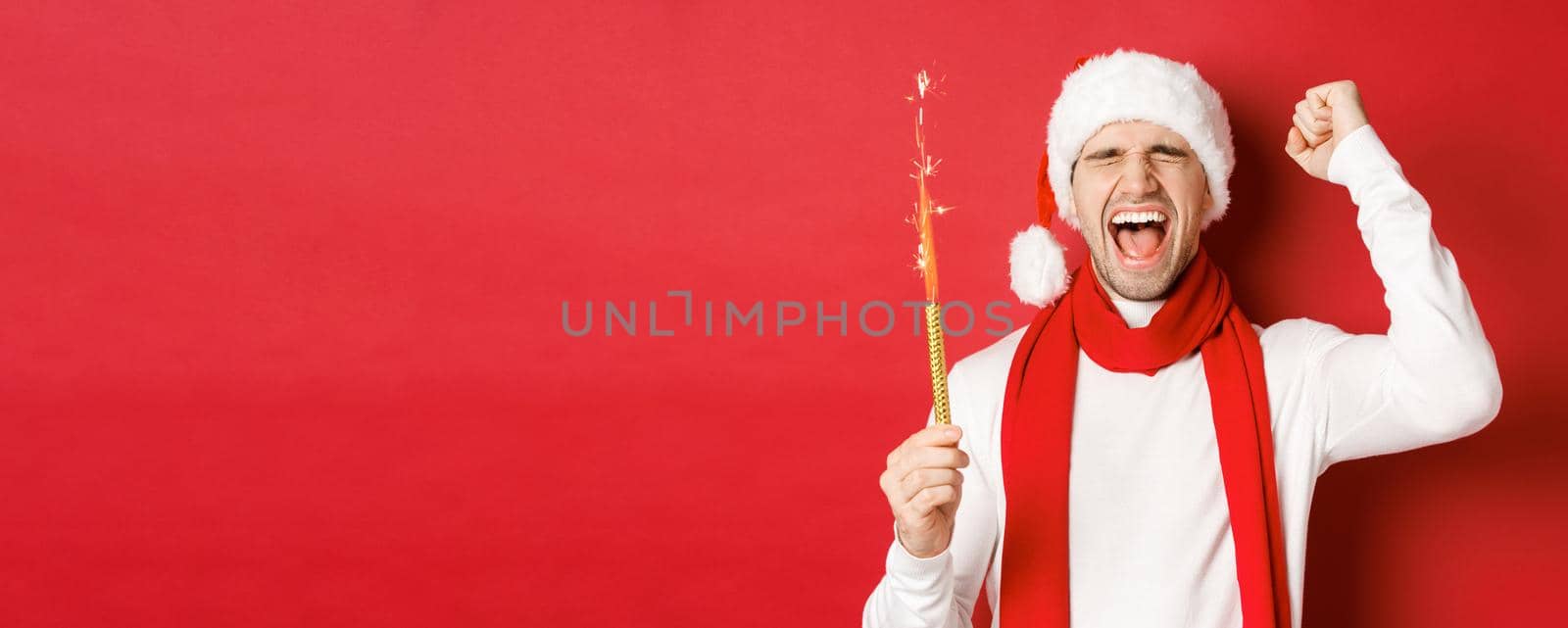 Concept of christmas, winter holidays and celebration. Handsome man celebrating new year and having fun, holding sparkler and smiling, wearing santa hat, standing over red background by Benzoix
