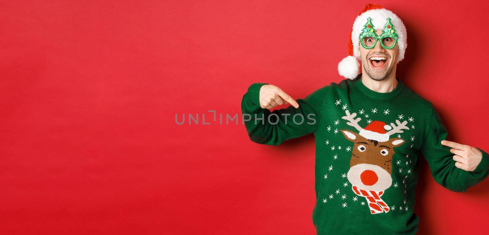 Image of happy man in party glasses and santa hat, pointing at his christmas sweater and smiling, standing over red background by Benzoix