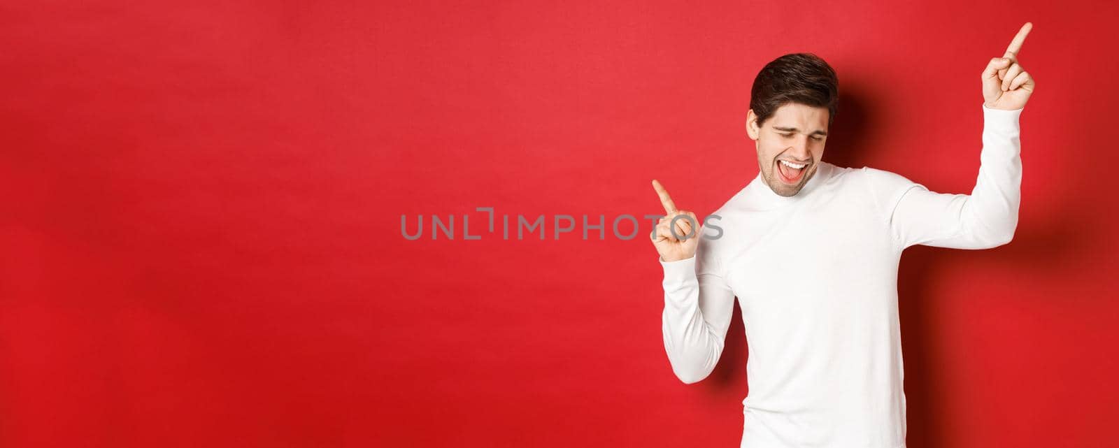Portrait of handsome man enjoying new year party, dancing and having fun, pointing fingers up, standing over red background.