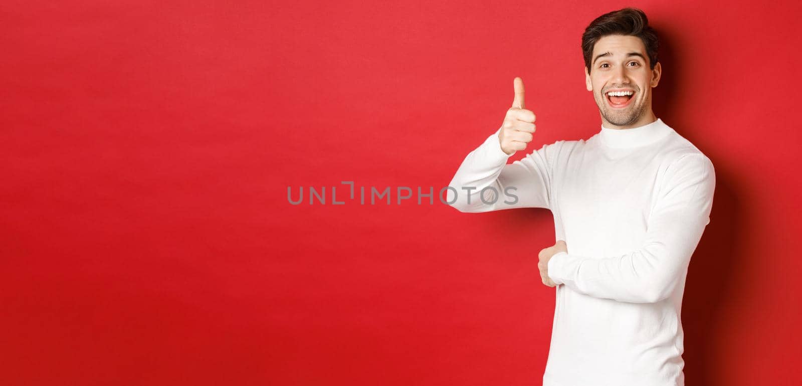 Concept of winter holidays, christmas and lifestyle. Portrait of cheerful, attractive man in white sweater, showing thumbs-up in approval, like good idea, standing over red background.