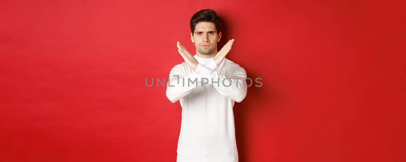 Image of serious and angry man in white sweater, express storng disapproval, showing cross sign to stop something bad, forbid action, standing over red background by Benzoix