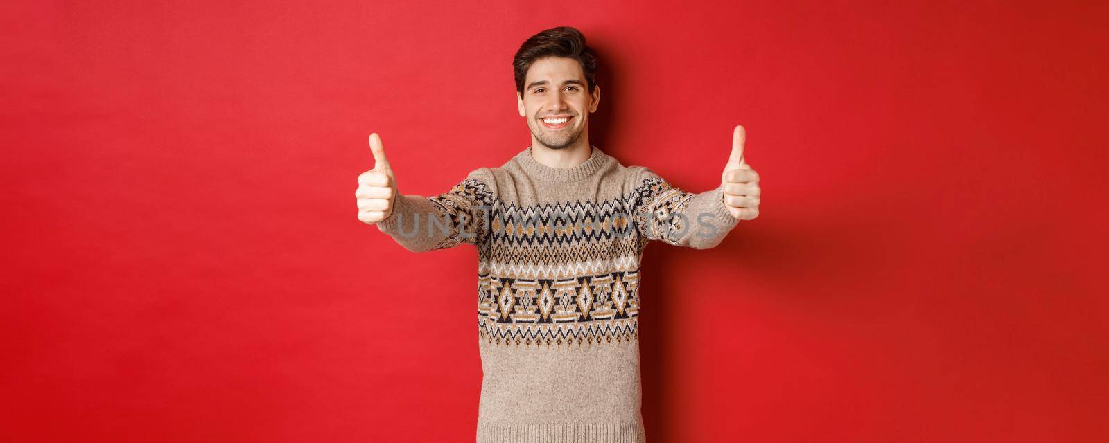 Image of handsome caucasian man in christmas sweater, showing thumbs-up in approval and smiling, wishing happy holidays, standing over red background by Benzoix