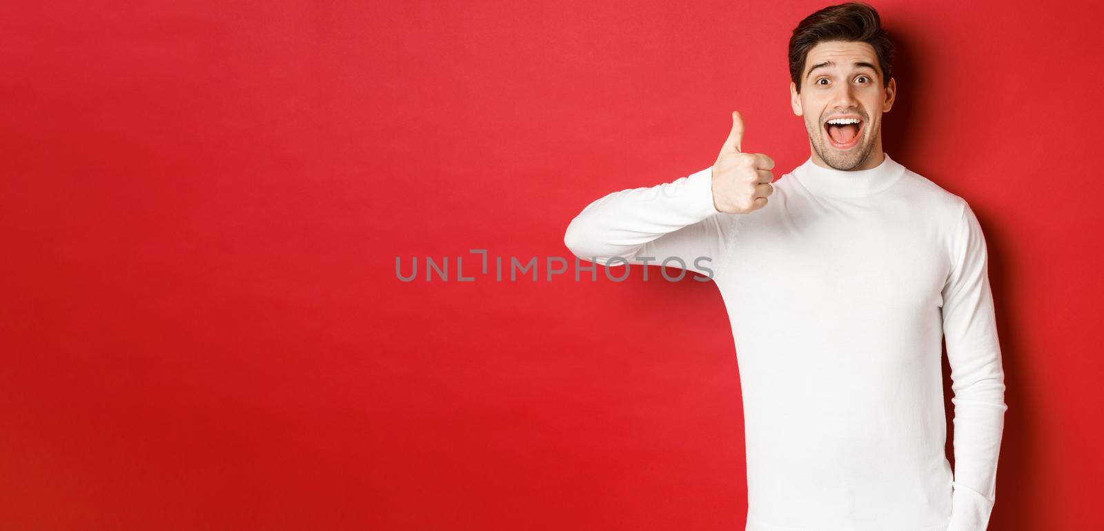 Concept of winter holidays, christmas and lifestyle. Excited good-looking man in white sweater, like good product, showing thumb-up in approval and smiling amazed, red background by Benzoix