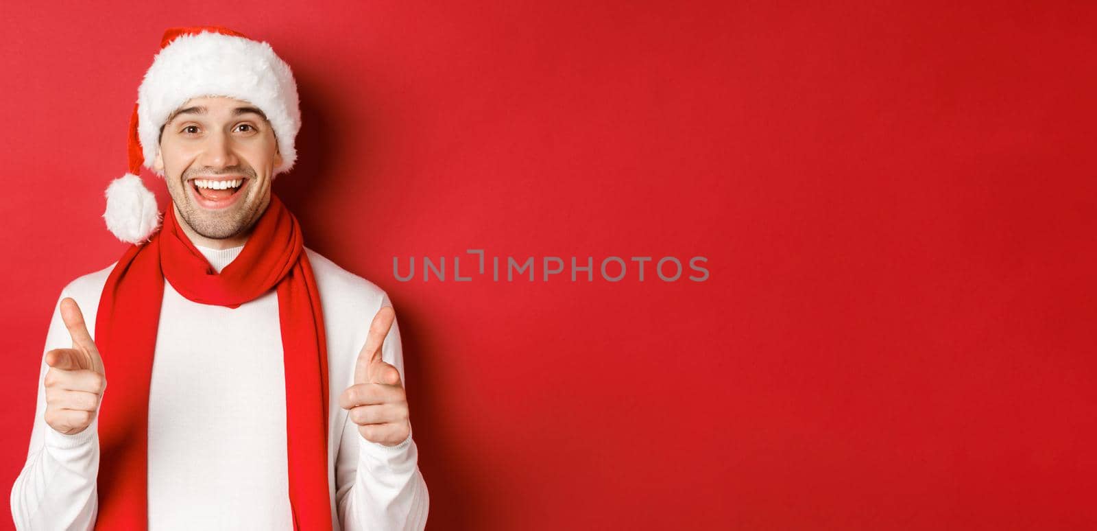 Concept of christmas, winter holidays and celebration. Cheeky man in santa hat and scarf, smiling and pointing fingers at camera, wishing happy new year, standing over red background by Benzoix