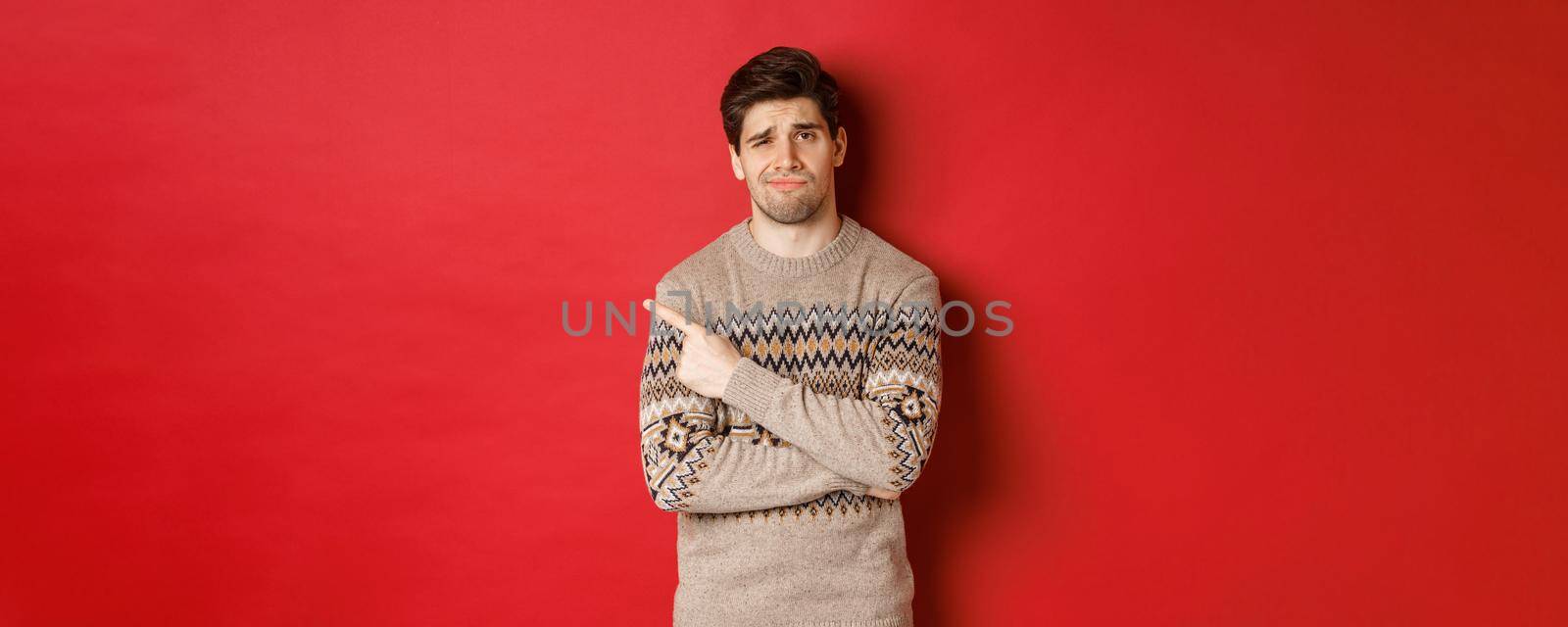 Image of skeptical and displeased man in christmas sweater, pointing at upper left corner unamused, showing bad xmas gift, standing over red background by Benzoix