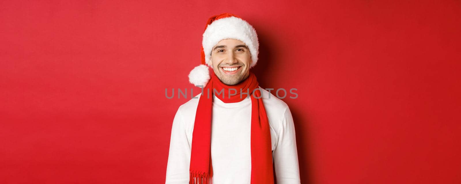 Concept of winter holidays, christmas and lifestyle. Close-up of handsome man with bristle, wearing christmas hat with scarf and smiling happy, wishing happy new year, red background.