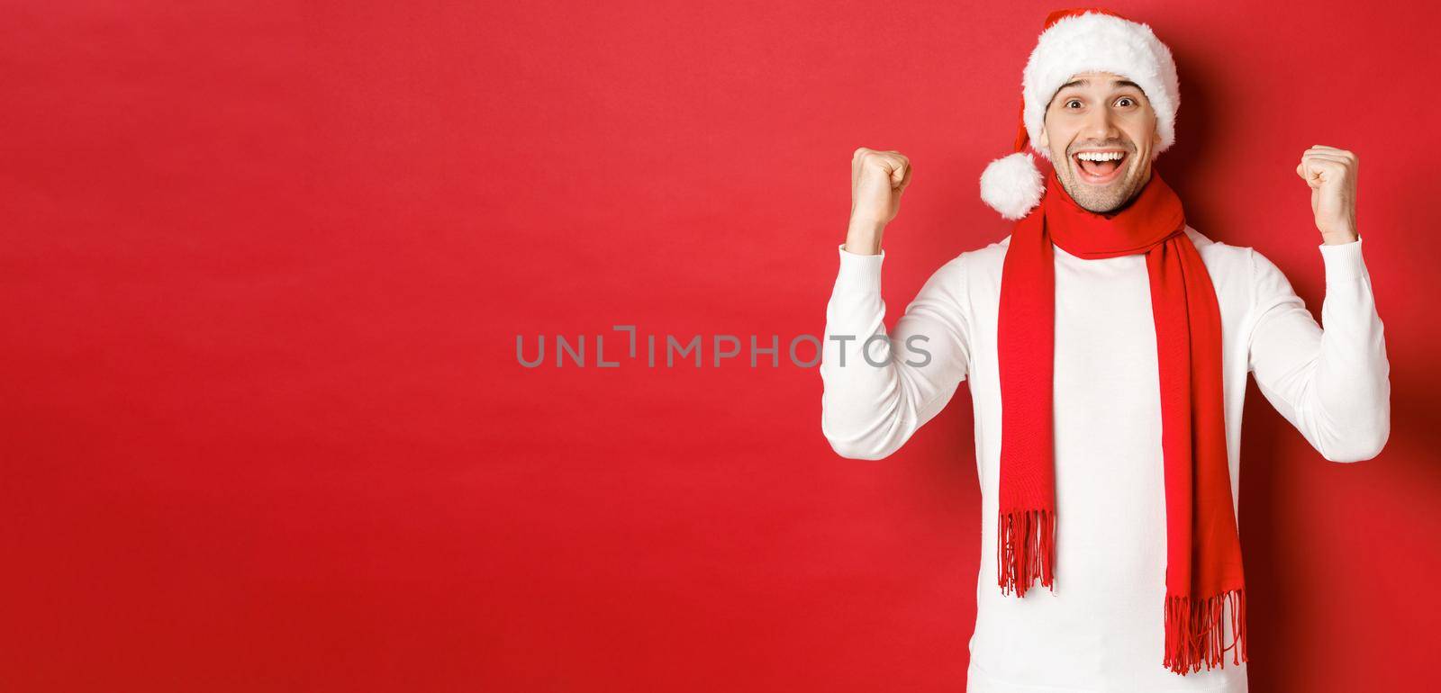 Portrait of happy and excited man in santa hat and scarf, rejoicing and winning something, celebrating new year, standing over red background.