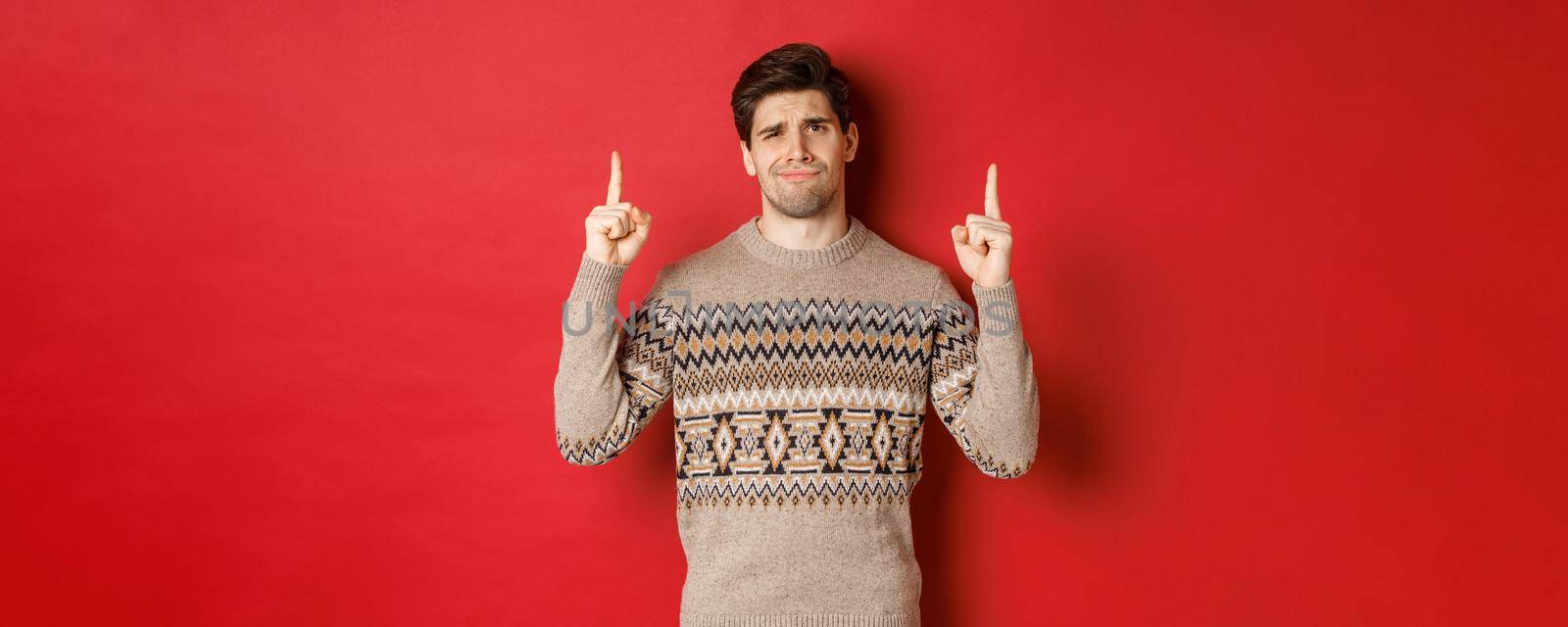 Image of skeptical and unamused handsome guy, showing bad christmas offer, pointing fingers up and grimacing displeased, express dislike, standing over red background by Benzoix