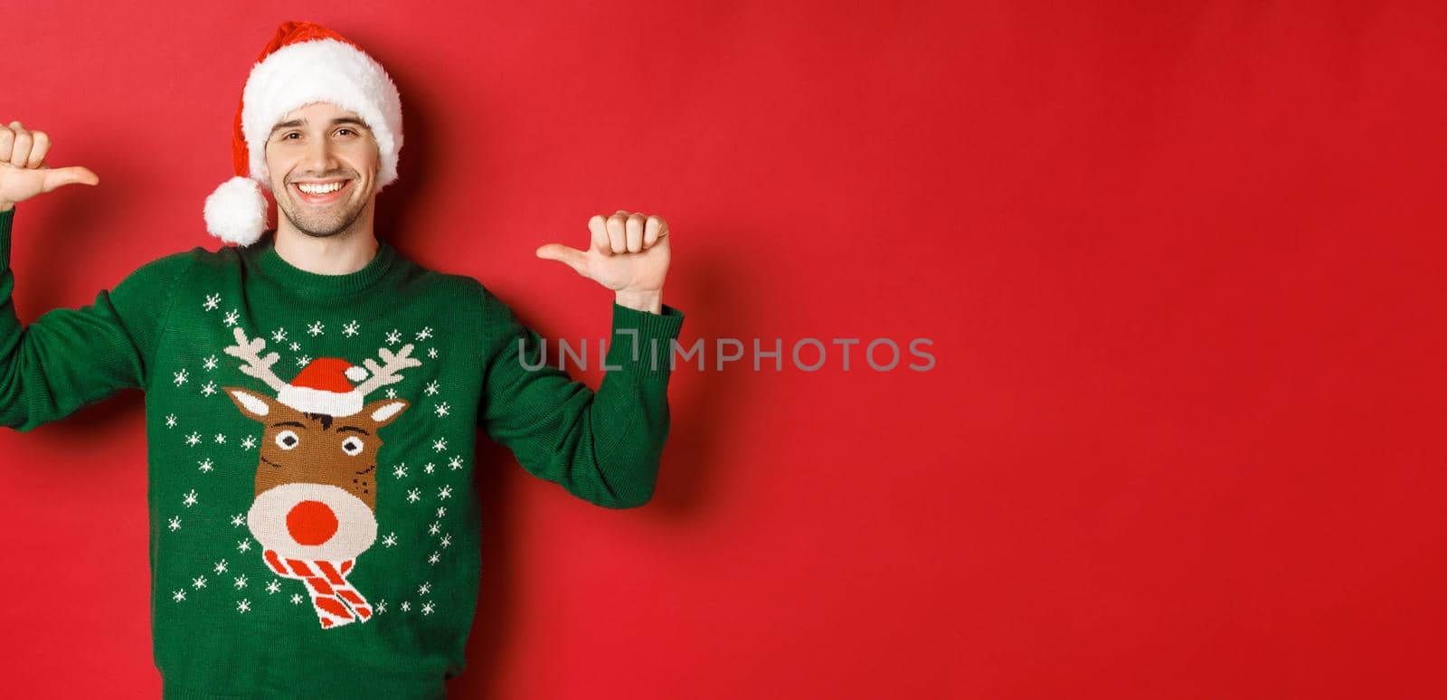 Concept of winter holidays, christmas and lifestyle. Happy attractive guy in santa hat and sweater, pointing at himself with pleased face, standing over red background.