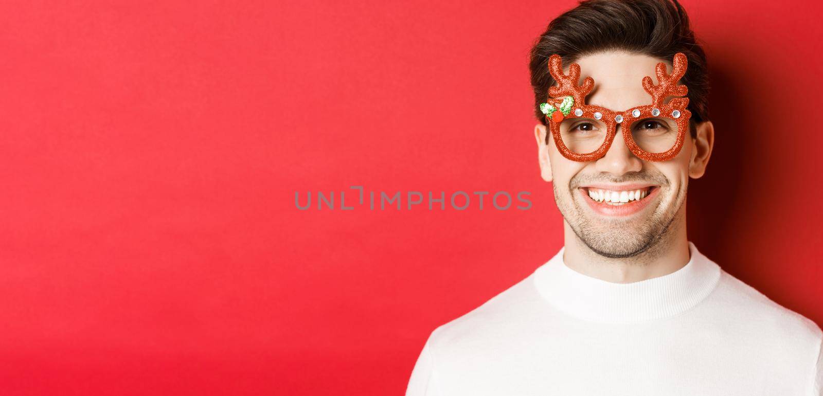 Concept of winter holidays, christmas and celebration. Close-up of attractive brunette guy in party glasses, smiling and looking happy, standing against red background.