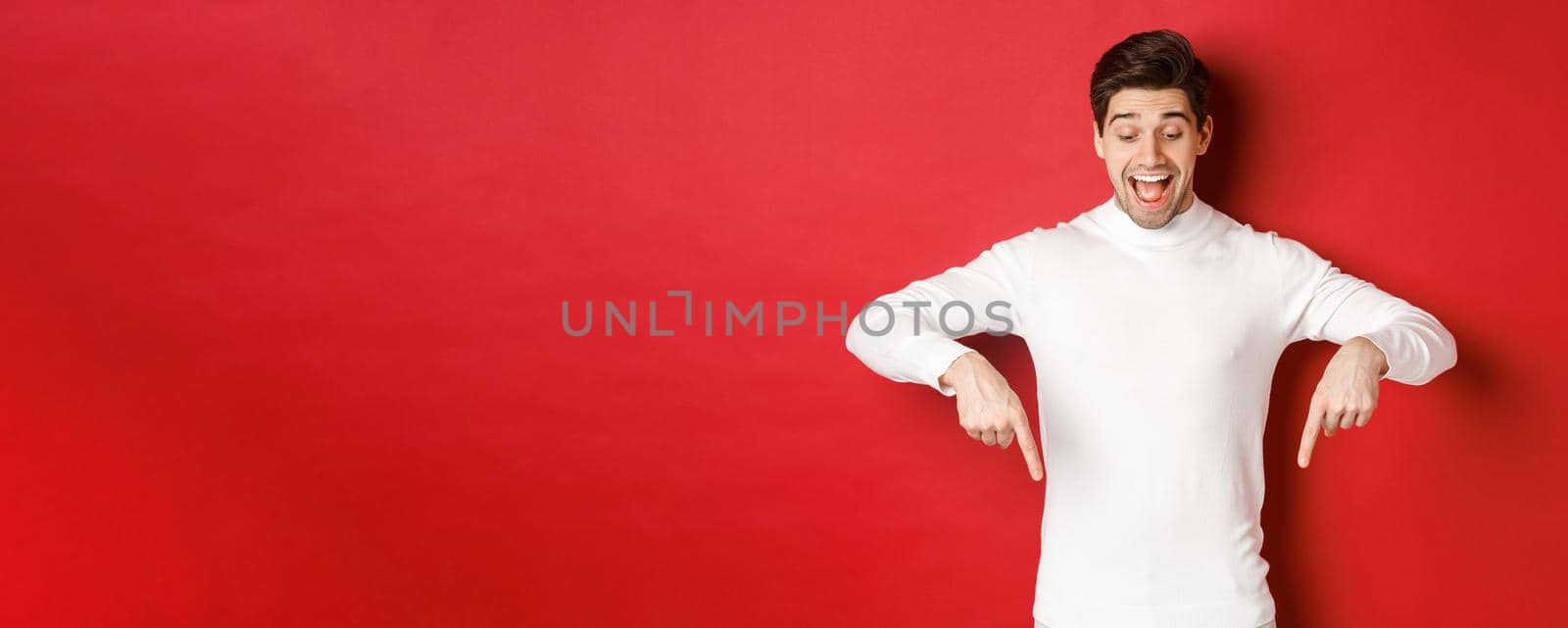 Portrait of happy good-looking man, pointing fingers down and looking at advertisement with excited smile, standing over red background.