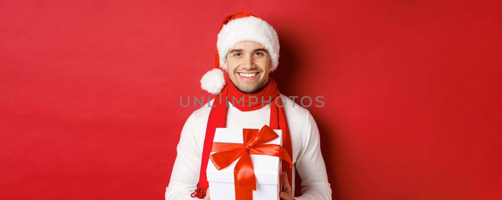 Concept of winter holidays, christmas and lifestyle. Close-up of attractive bearded guy in santa hat and scarf, holding a present, giving new year gift and smiling, red background.