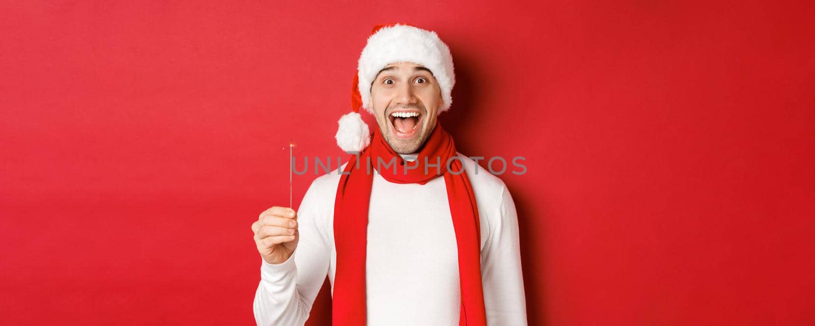 Concept of christmas, winter holidays and celebration. Close-up of happy handsome man in santa hat and scarf, having fun on new year party, holding sparkler and smiling, red background.