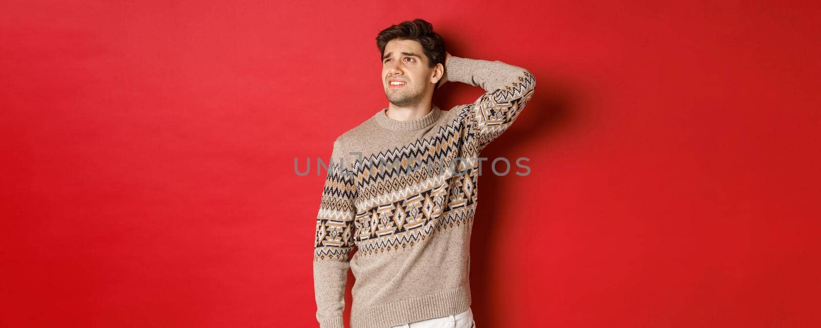 Image of puzzled and indecisive man in christmas sweater, looking at upper left corner, thinking about new year holidays gifts, standing over red background.
