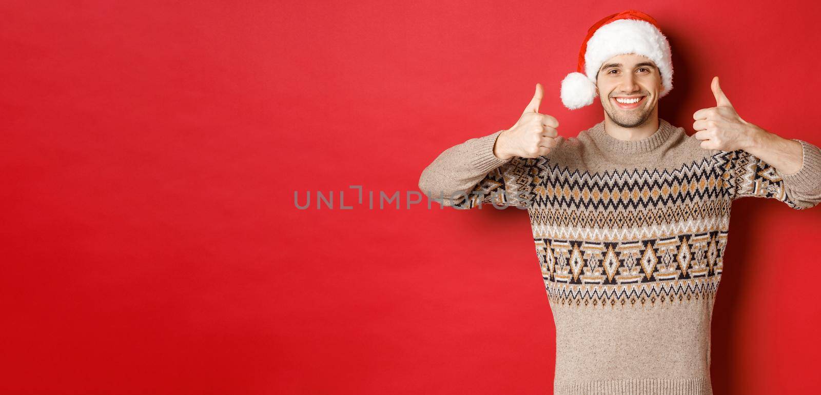 Portrait of attractive happy man in santa hat and sweater, showing thumbs-up in approval and smiling, wishing merry christmas, standing over red background by Benzoix