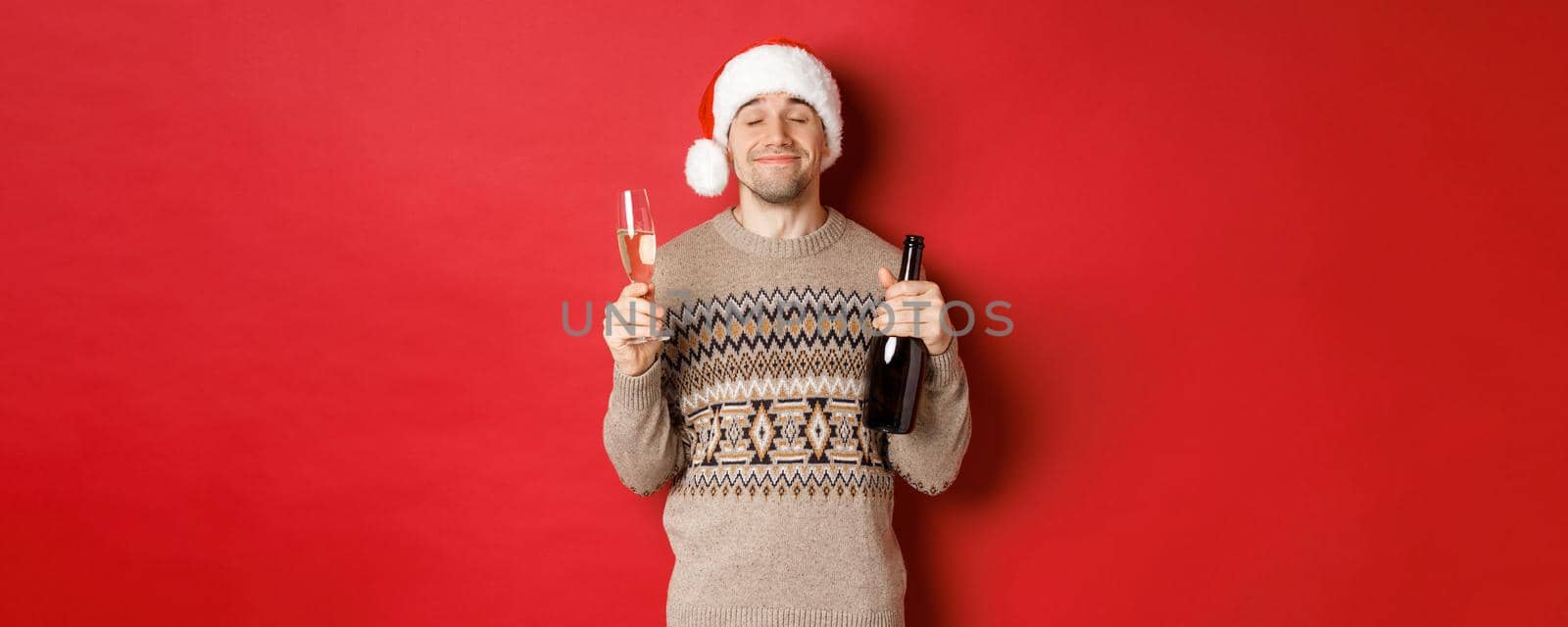 Concept of winter holidays, christmas and celebration. Image of pleased smiling man in santa hat and sweater, drinking on new year, holding bottle of champagne and filled glass, red background.