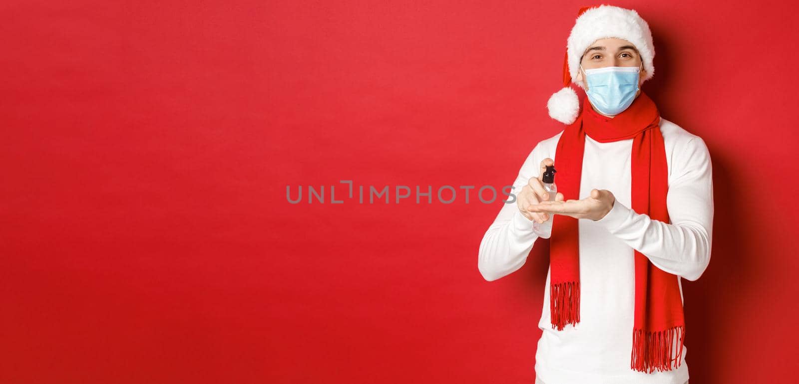 Concept of covid-19, christmas and holidays during pandemic. Happy young man in santa hat and medical mask, sanitize hands with antiseptic and smiling, standing over red background.