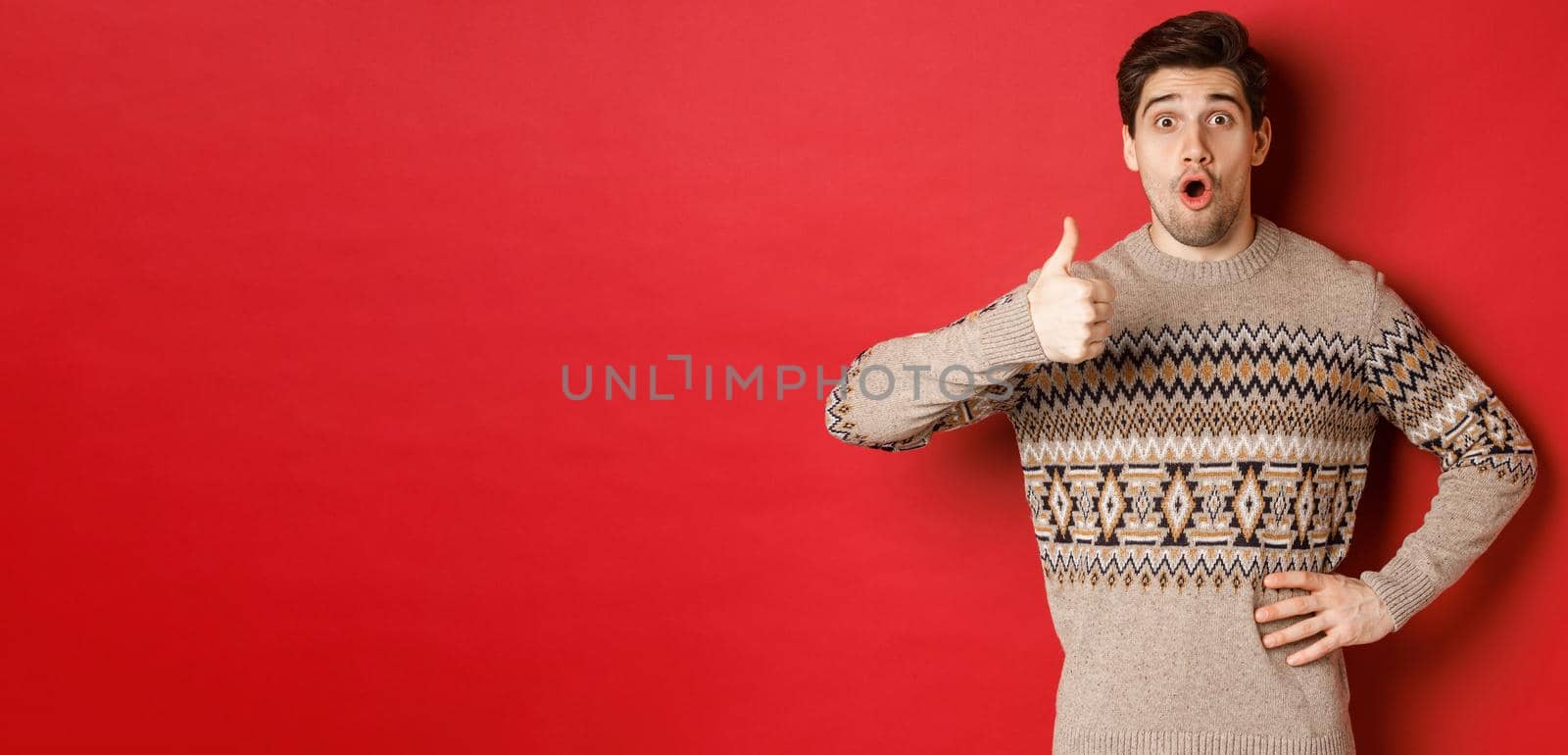 Image of handsome caucasian man in christmas sweater looking amazed, showing thumb-up in approval, like something, standing over red background by Benzoix
