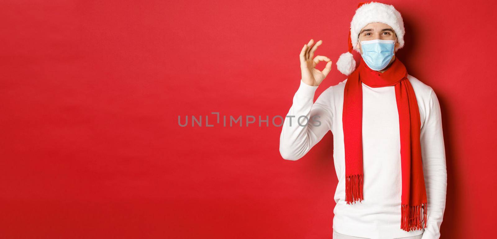 Concept of covid-19, christmas and holidays during pandemic. Portrait of happy and satisfied man in medical mask and santa hat, showing okay sign in approval, recommending something.