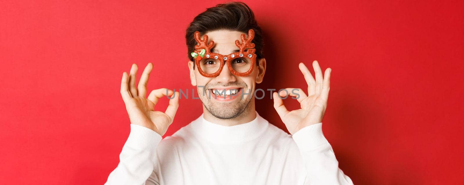 Concept of winter holidays, christmas and celebration. Close-up of attractive man in party glasses, showing okay signs and smiling, praise something good, red background.