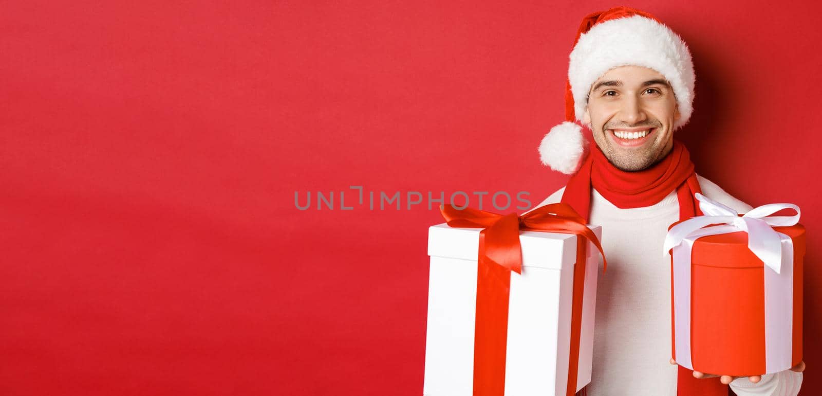 Concept of winter holidays, christmas and lifestyle. Close-up of smiling handsome guy in santa hat and scarf, wishing happy new year and holding gifts, standing with presents over red background.