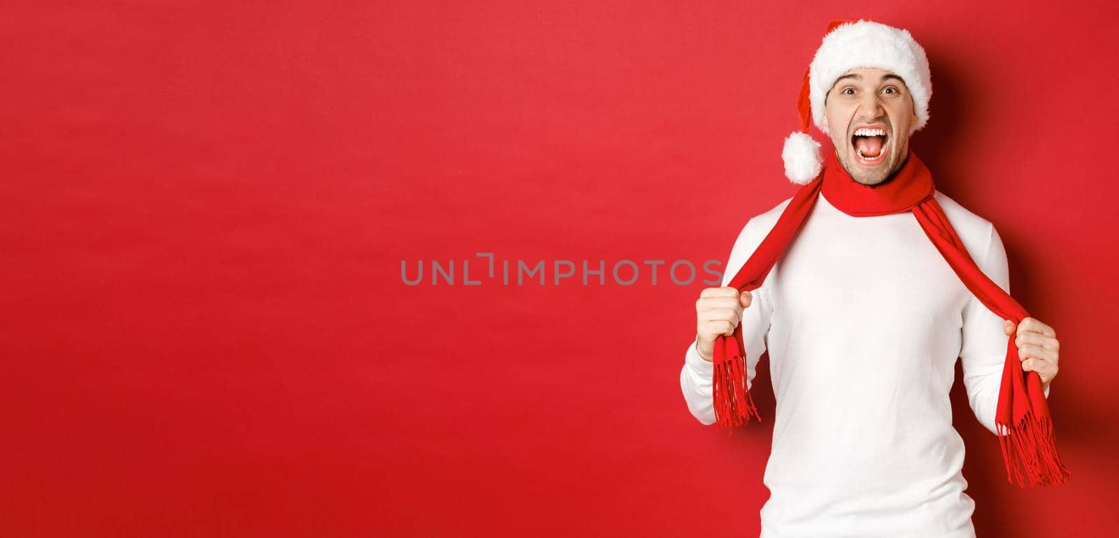 Image of angry adult man hate christmas, wearing scarf and santa hat, yelling distressed, standing mad over red background by Benzoix