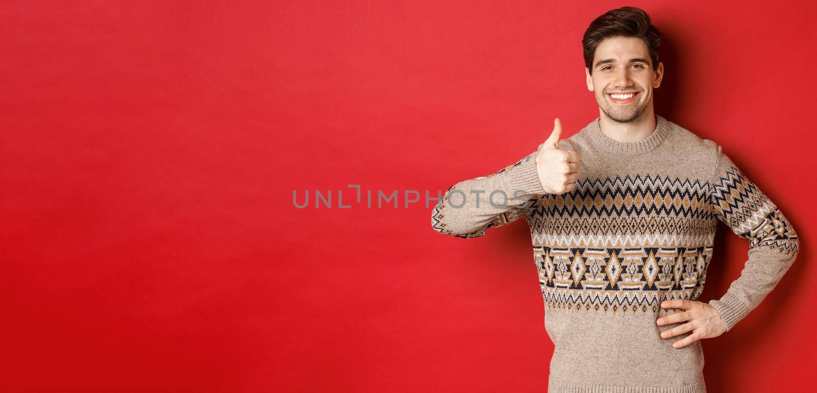 Portrait of happy handsome man in christmas sweater, smiling and looking satisfied, showing thumbs-up in approval, like new year holiday, standing over red background.