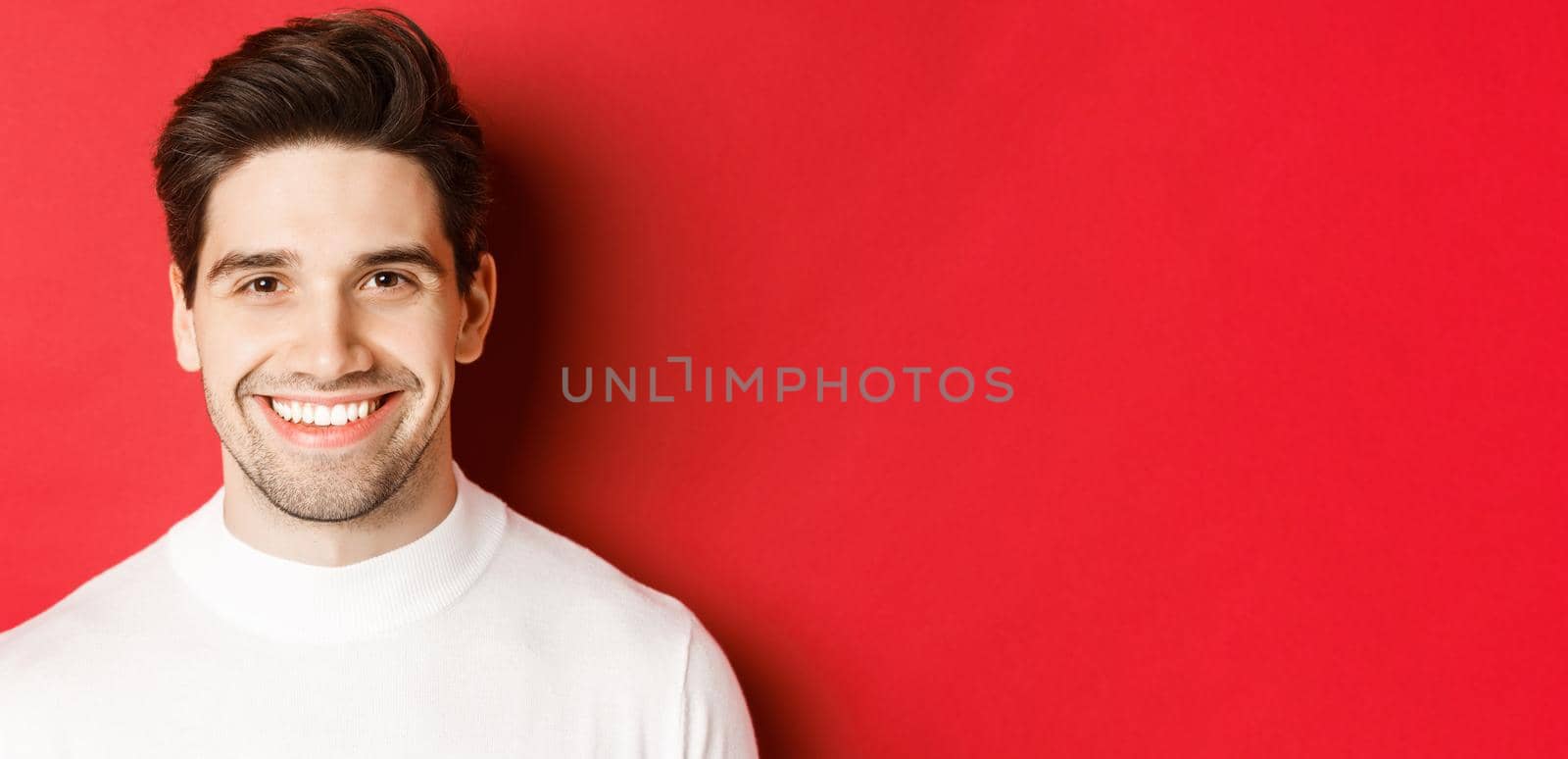 Close-up of handsome smiling brunette man, wearing white sweater, smiling happy and confident, standing against red background by Benzoix