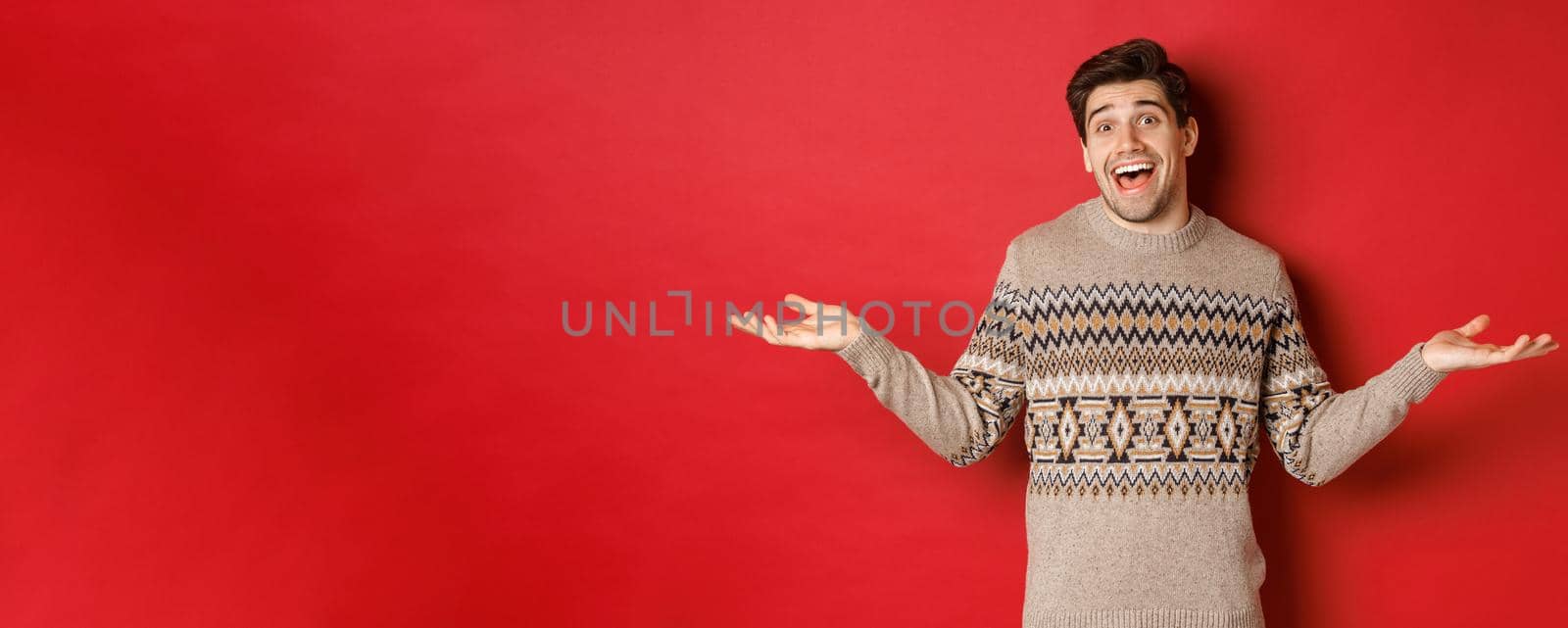 Portrait of happy and surprised, handsome caucasian guy, wearing christmas sweater, spread hands sideways and looking clueless, standing over red background.
