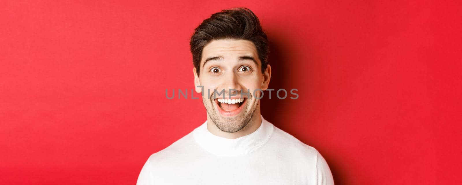 Close-up of surprised happy guy in white sweater, looking amused, hear interesting news, standing over red background.