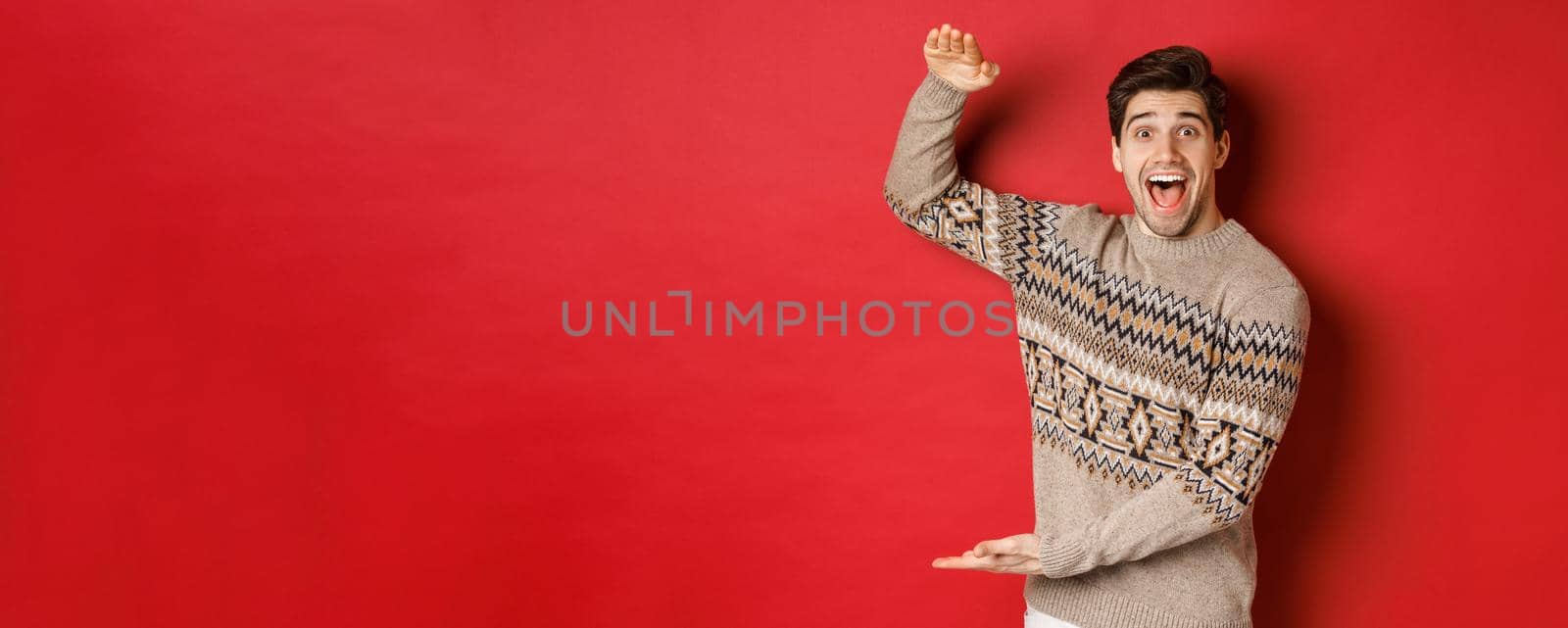 Portrait of excited and happy handsome man in christmas sweater, showing something big, holding large gift for holidays, standing over red background.