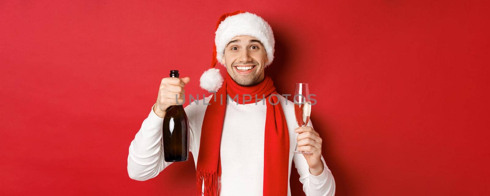 Concept of winter holidays, christmas and lifestyle. Close-up of cheerful handsome man, holding champagne bottle and glass, making toast for new year and celebrating, red background.