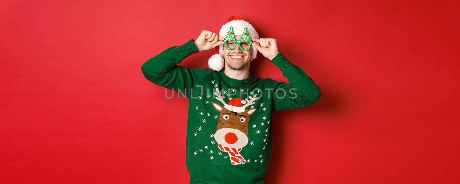 Portrait of attractive smiling man in santa hat, party glasses and sweater, celebrating new year holidays, standing against red background by Benzoix