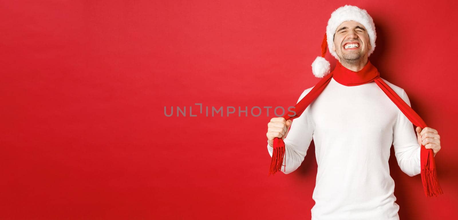 Image of distressed and frustrated man in santa hat, chocking himself with scarf from sadness, standing against red background.