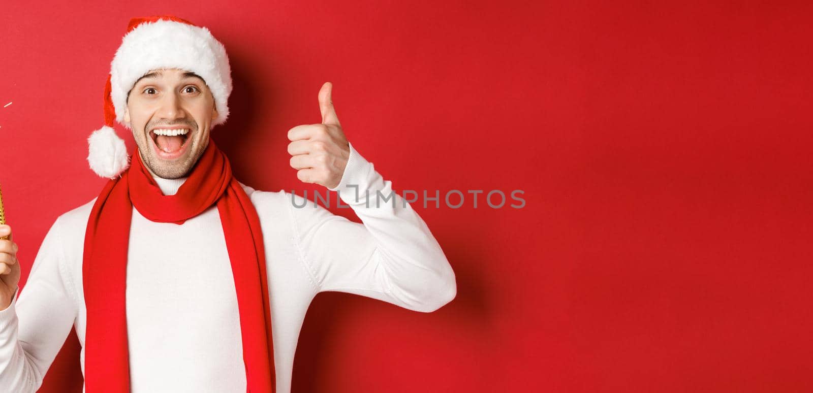 Concept of christmas, winter holidays and celebration. Handsome man celebrating new year and having fun, holding sparkler and showing thumb-up, standing over red background.