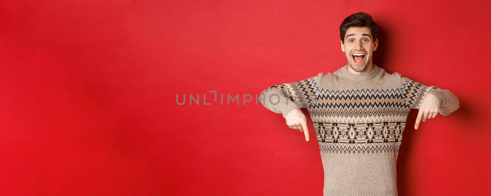 Concept of christmas celebration, winter holidays and lifestyle. Excited caucasian male model in xmas sweater, pointing fingers down and making announcement, showing advertisement, red background.