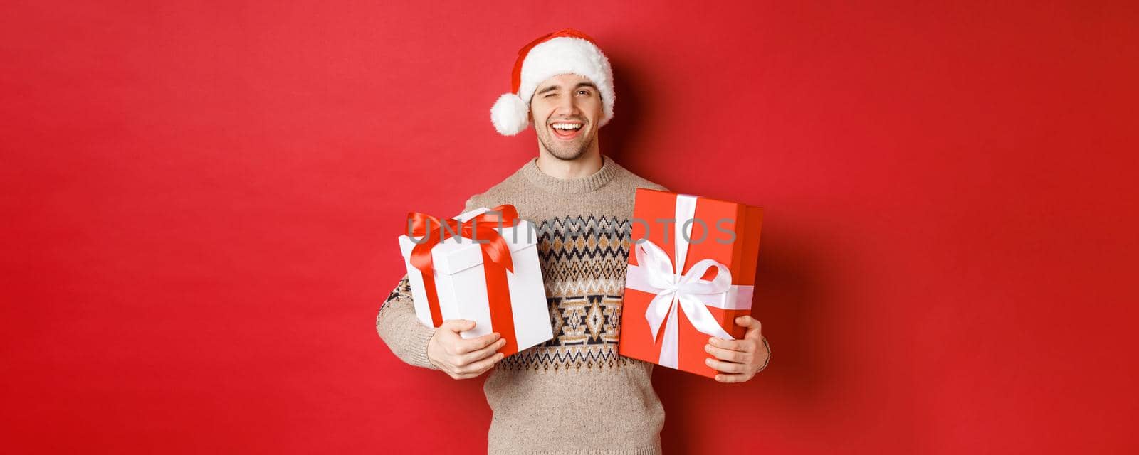 Concept of winter holidays, new year and celebration. Portrait of confident and cheeky young man prepared gifts for christmas, winking and holding presents, standing over red background by Benzoix