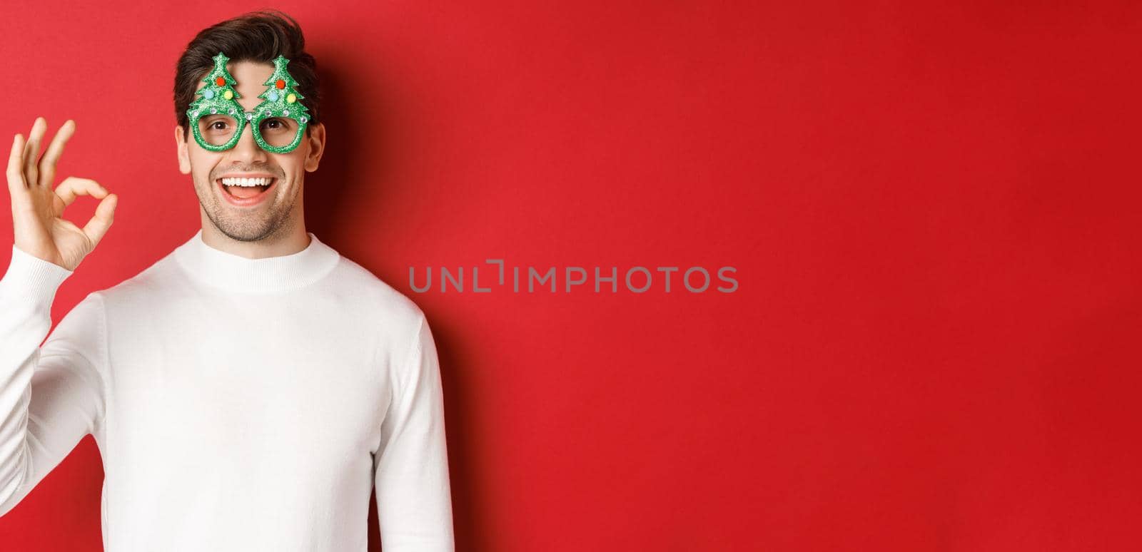 Happy caucasian guy wishing merry christmas, celebrating winter holidays, showing okay sign and smiling pleased, standing over red background by Benzoix