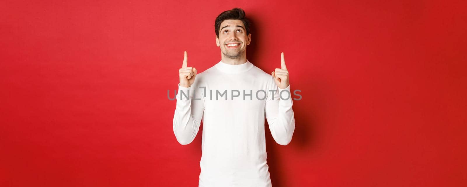 Concept of winter holidays. Image of happy attractive man in white sweater, smiling while pointing and looking up at copy space on red background.